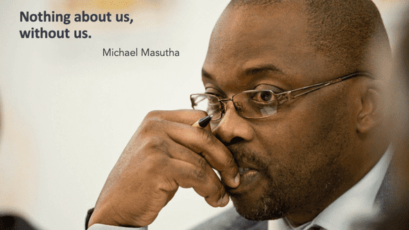 Photo of Michael Masutha, in profile position to the right of the photo. He wears a suit and glasses, and holds a pen in his hand, held to his mouth, while he gazes into the distance. In the top left corner of the image is a quote attributed to Mr. Masutha that reads 'Nothing about us, without us'